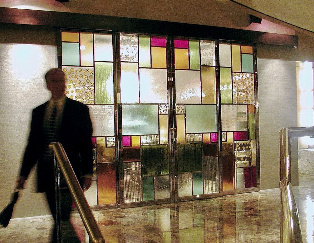 Man walks past colorful cast glass panels by a stairwell, with gold-toned metal railing.