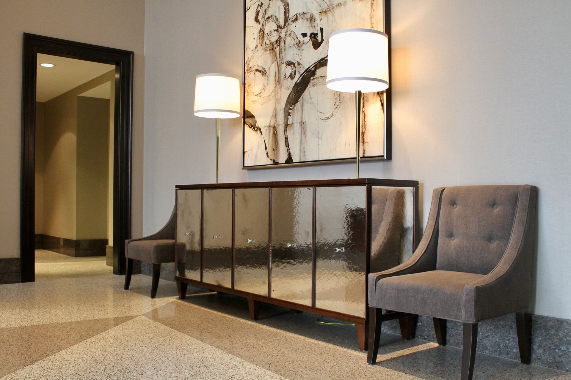 A foyer with a mirrored console table, two gray tufted chairs, two lamps, and abstract wall art on a light gray wall.