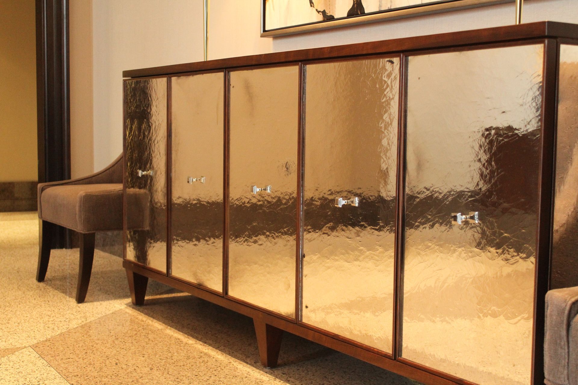 An antique mirrored cabinet is sitting next to a chair in a hallway.