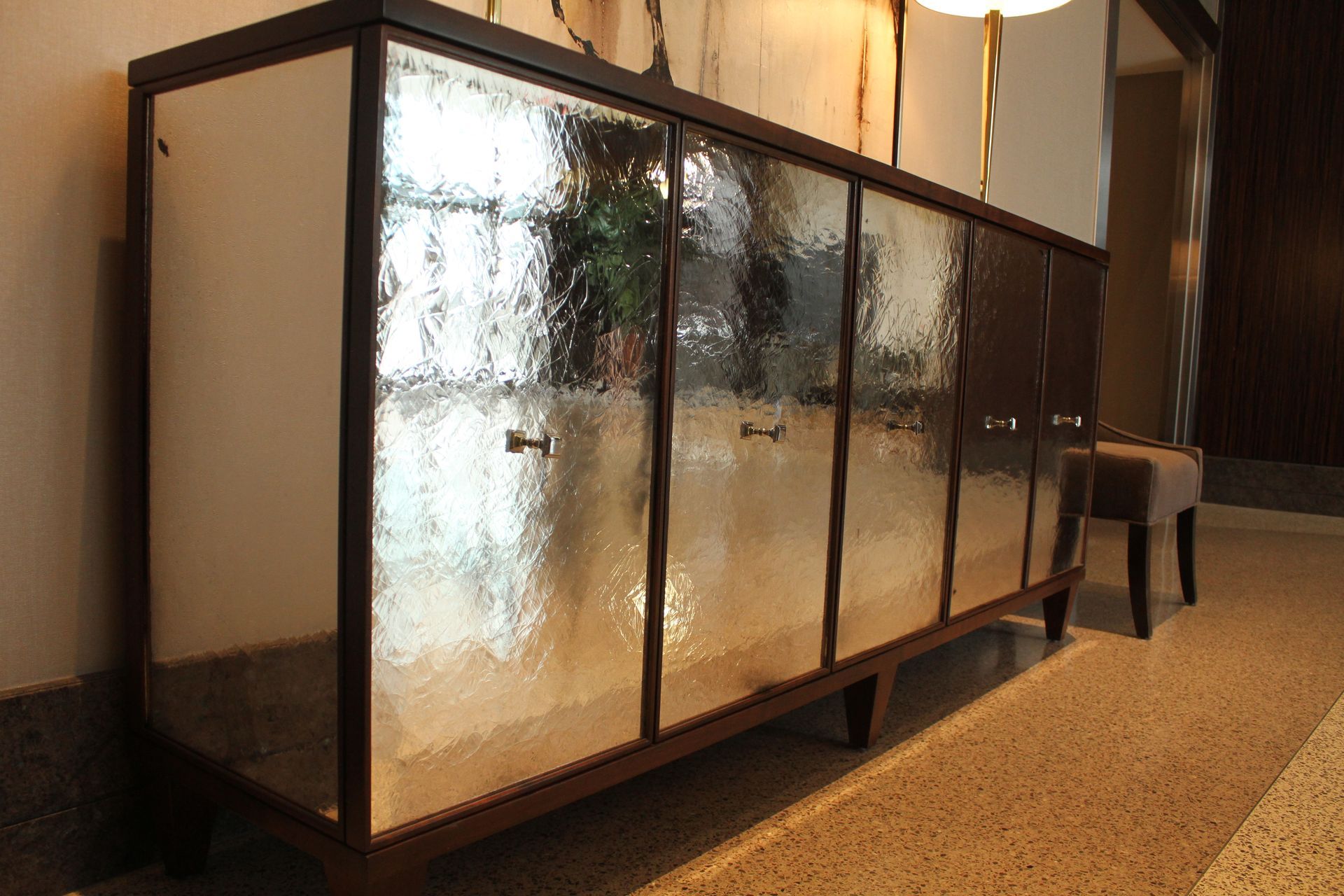 Dark-framed cabinet with textured glass doors, on a speckled floor, with a small stool.