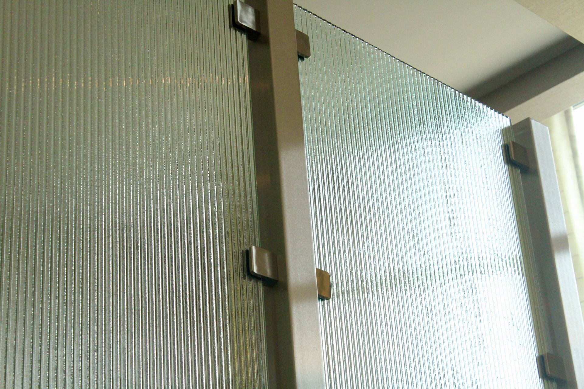 A close-up of textured glass panels held by a central metal post and small brackets in a modern interior setting.