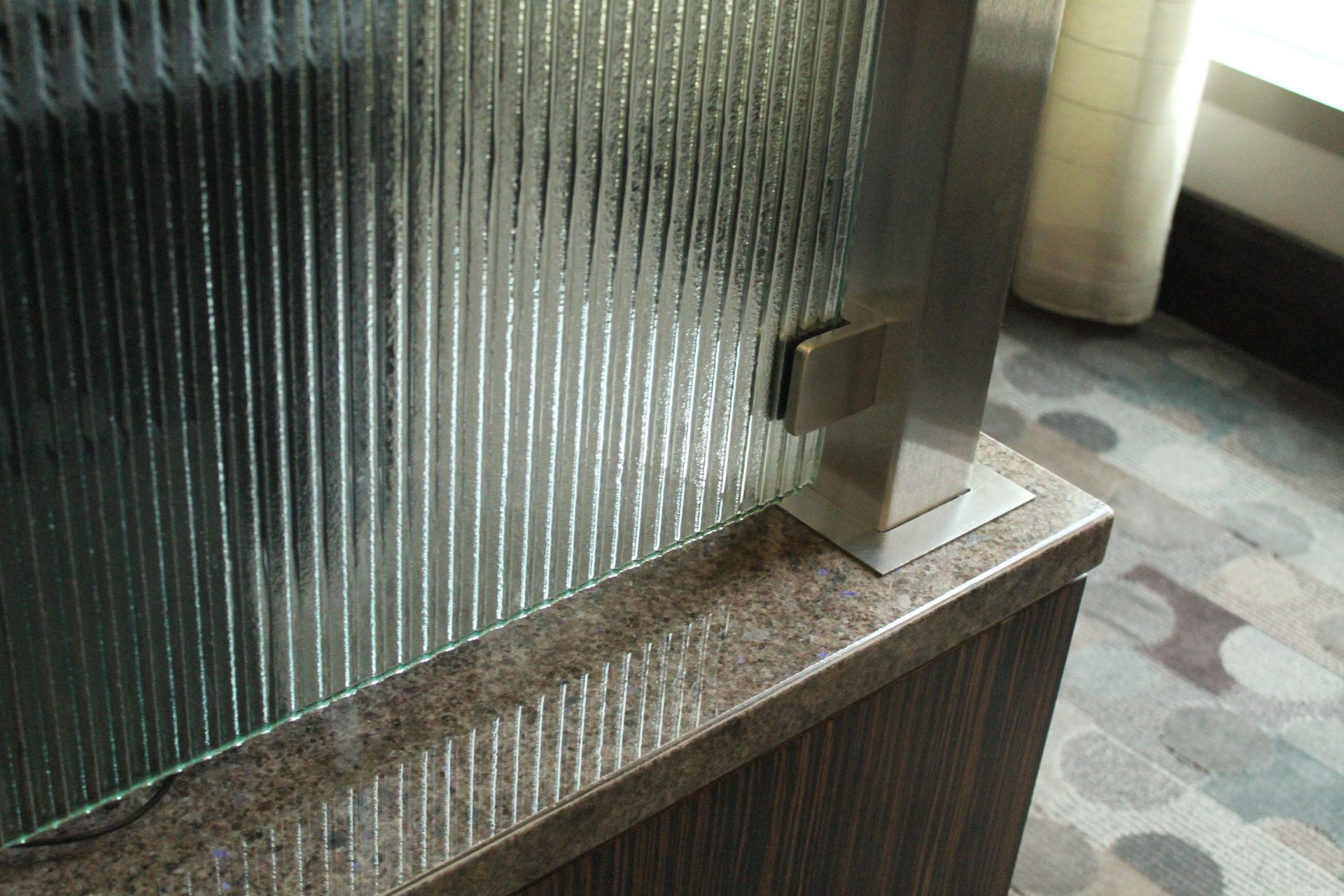 A vertical, ribbed glass partition panel mounted with a brushed metal bracket to a granite-topped wooden counter.
