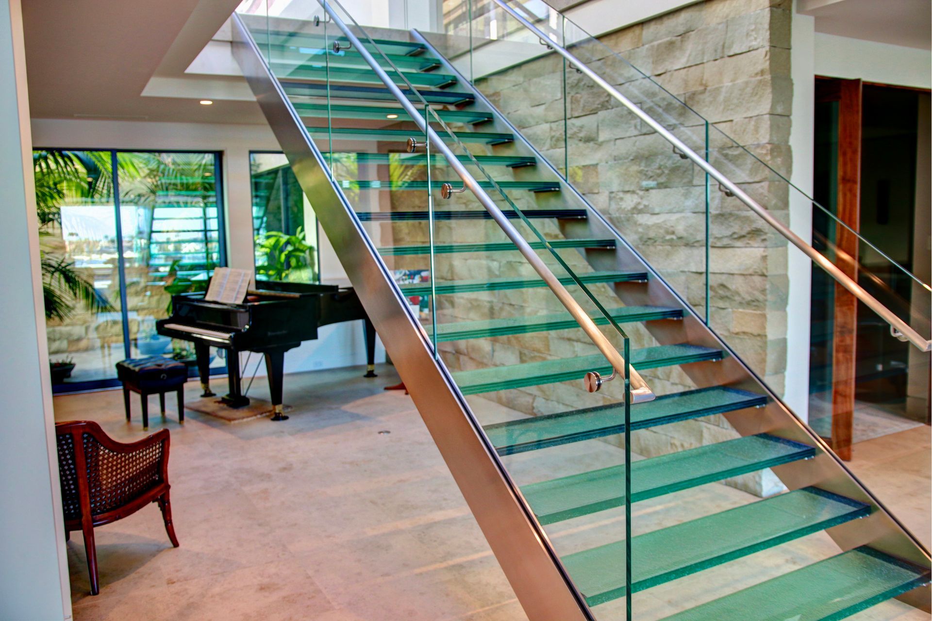 Modern indoor staircase with glass treads and metal railings, featuring a grand piano and seating in the background.