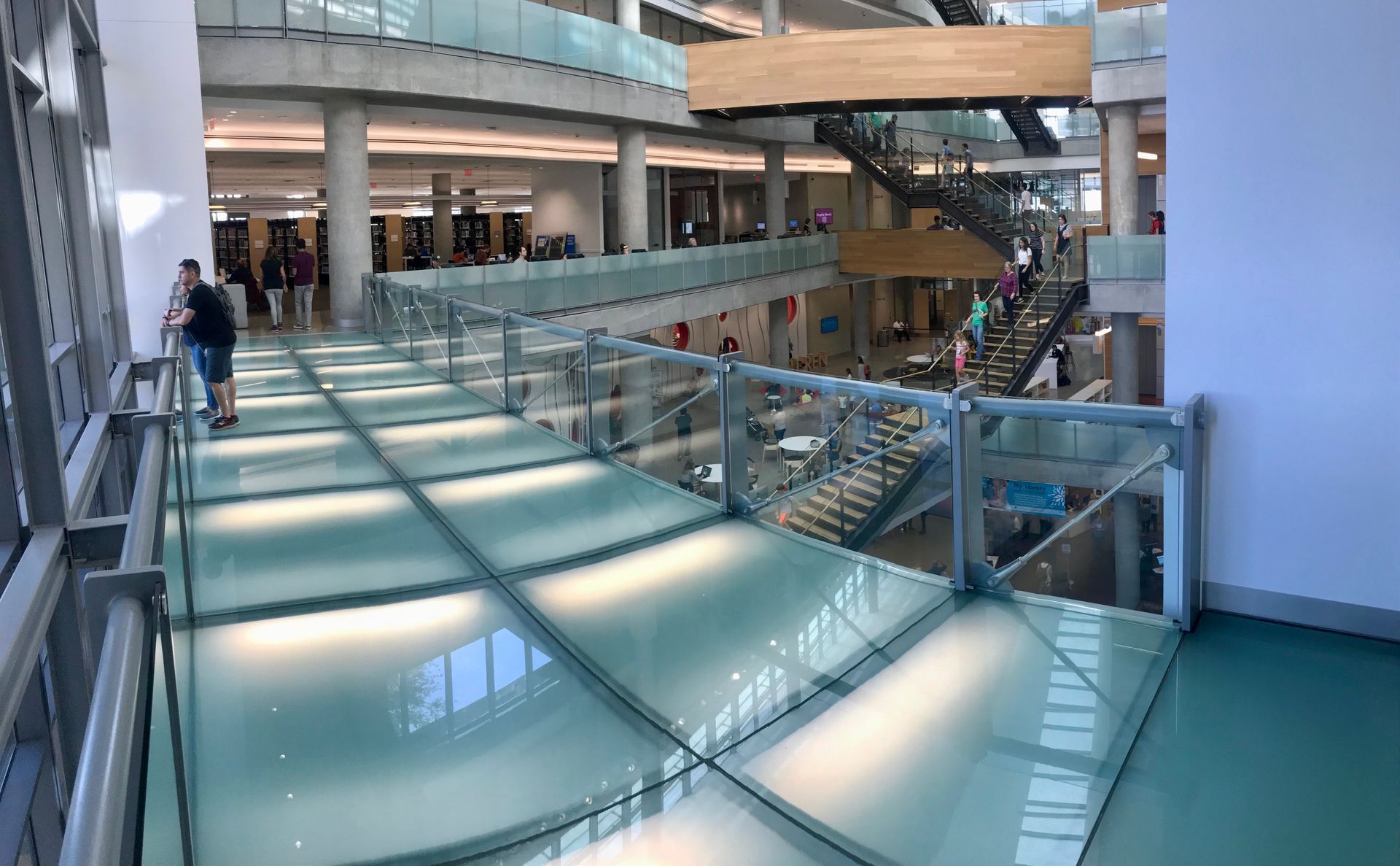 A person stands on a glass-floored walkway overlooking an open, multi-story library atrium with stairs and balconies.