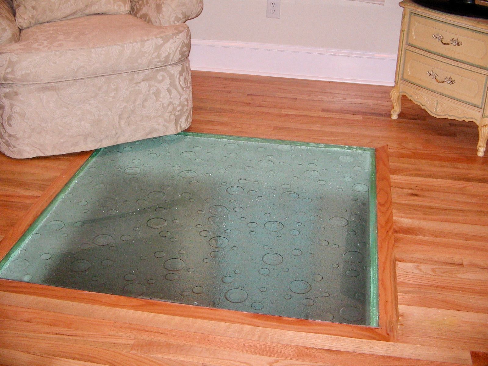 Glass-covered opening in wooden floor; armchair and nightstand in the background.