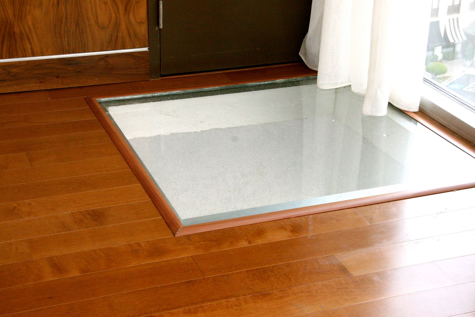 Glass floor panel inset in wooden floor near a window and dark door.