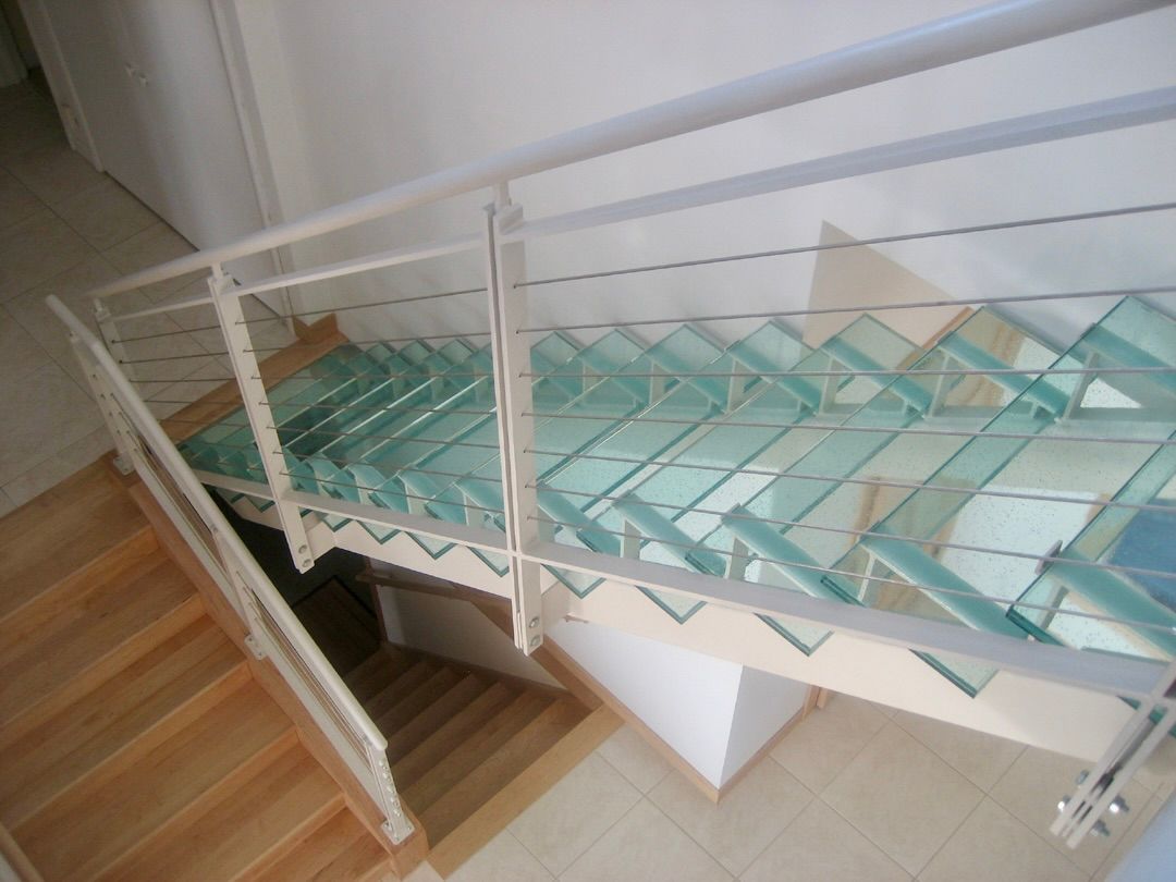 A high-angle view of a split staircase with transparent glass treads, light metal railings, and a wooden lower landing.