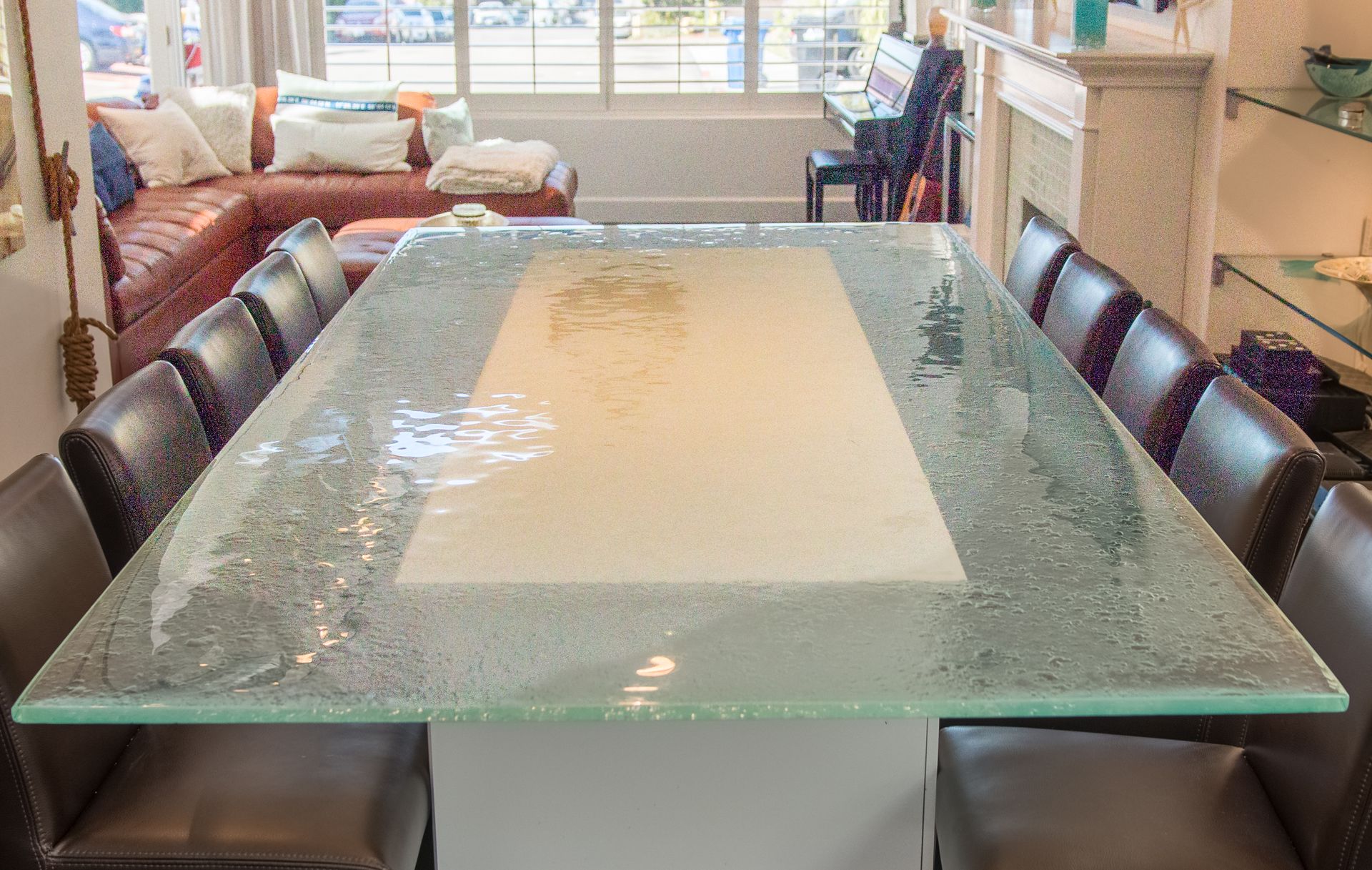 A dining room features a long table with a glass top and a central stone inset, surrounded by dark brown leather chairs.