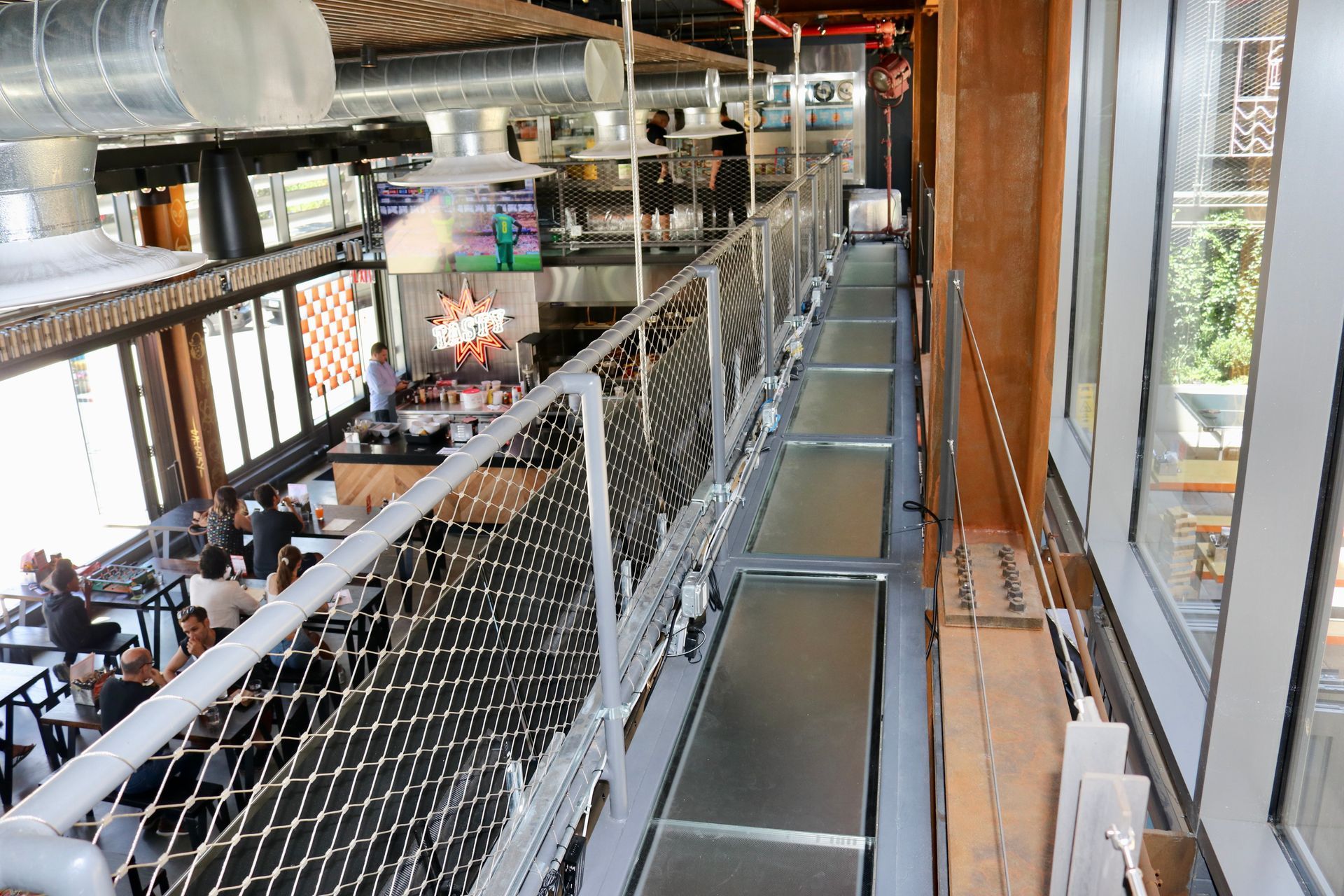 An elevated glass catwalk with a safety net overlooking a busy restaurant with bar seating and an open kitchen.