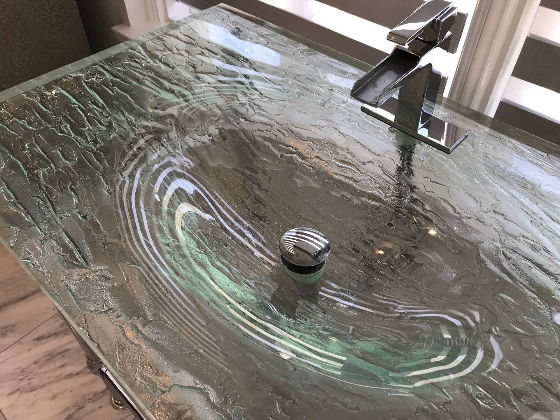 Glass countertop sink with swirling patterns, chrome faucet, and drain stopper.