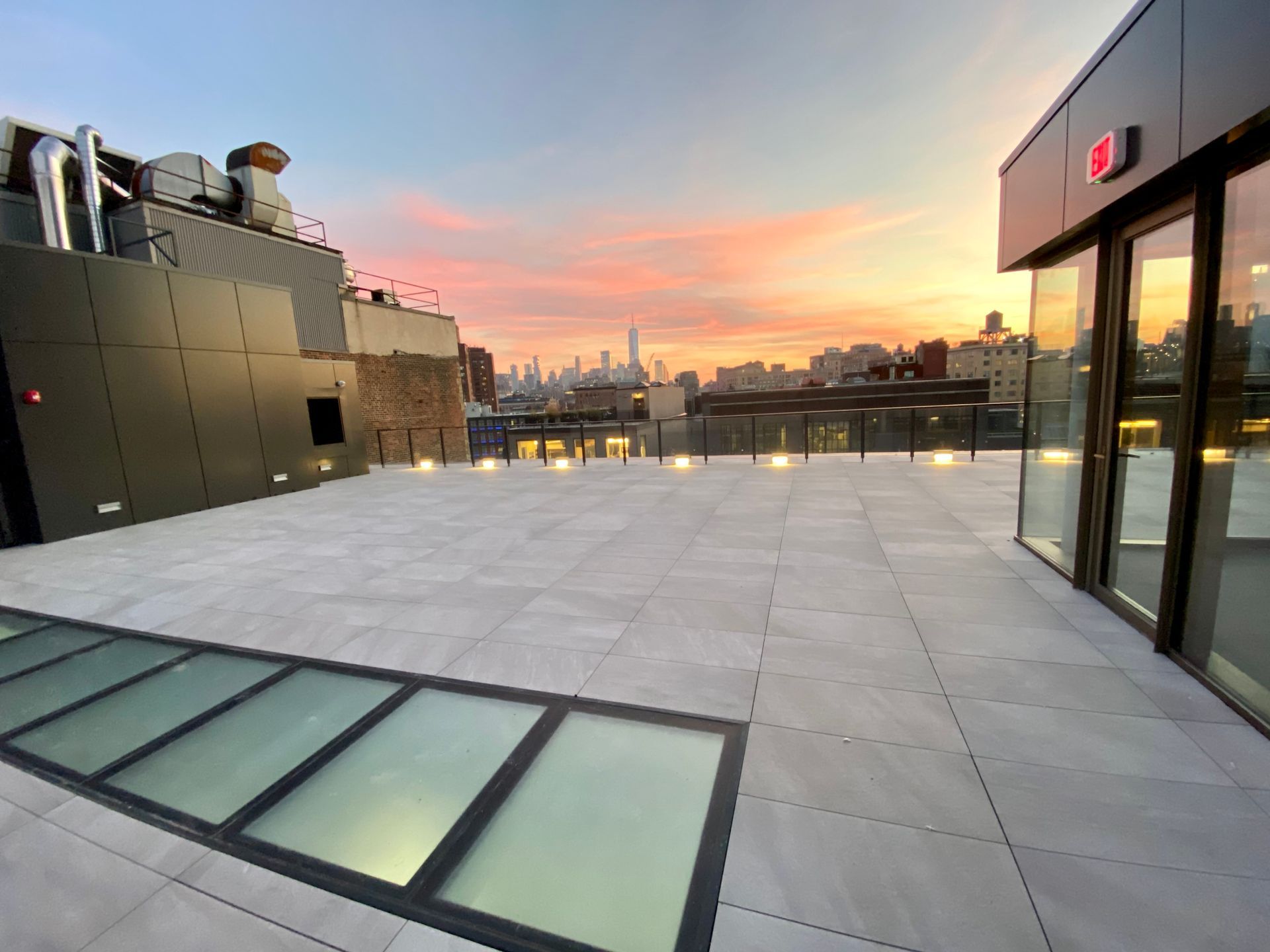 Rooftop terrace with light stone tiles, a glass floor inlay, and city skyline view during a vibrant sunset.