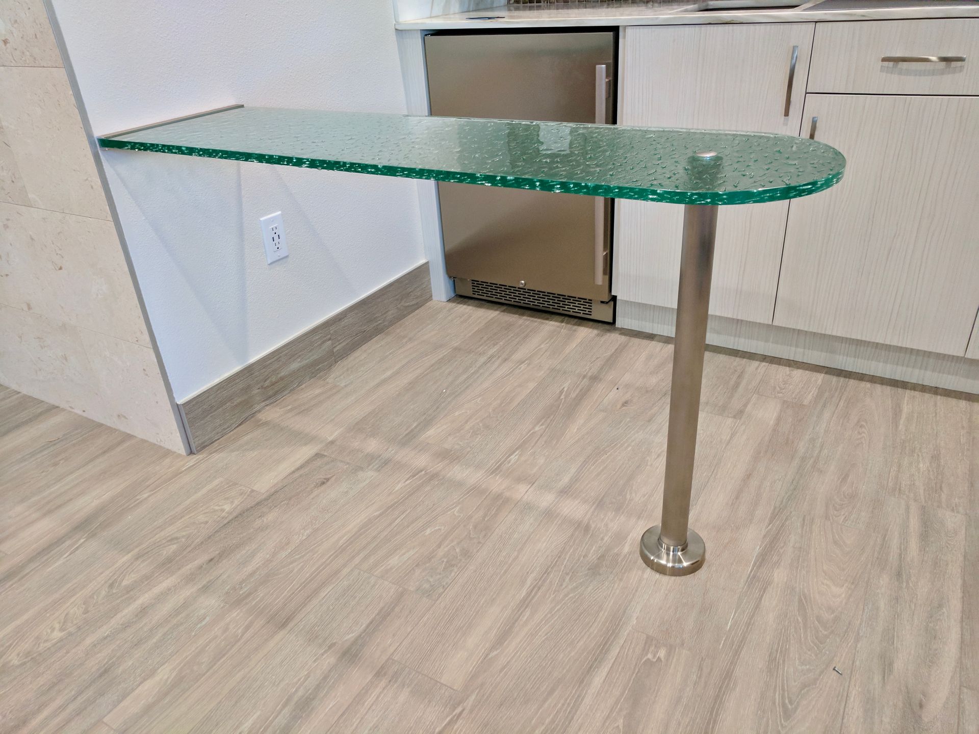 Glass countertop table, one metal leg, attached to a white wall. Stainless steel appliances in background.