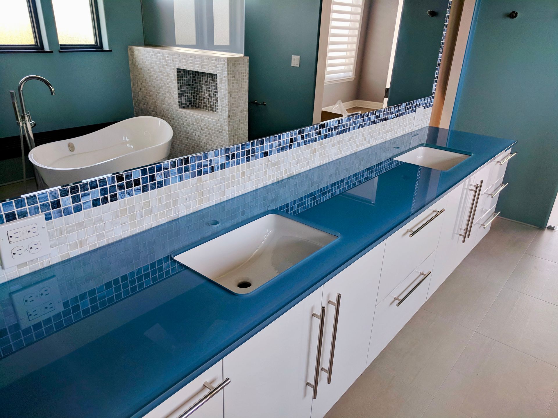 Blue countertop with white cabinets and sinks in a bathroom with a tub and mosaic wall.