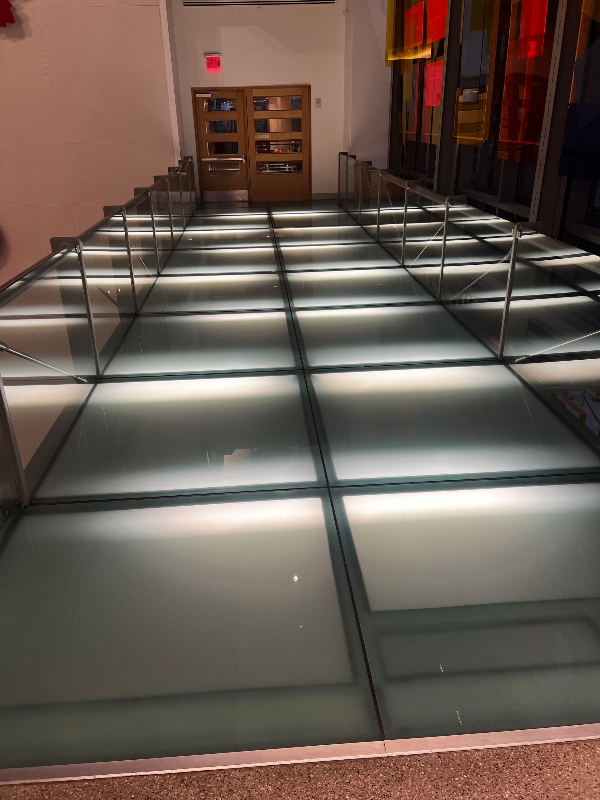 Glass-floored walkway with lights, leading to a door. Handrails on either side.