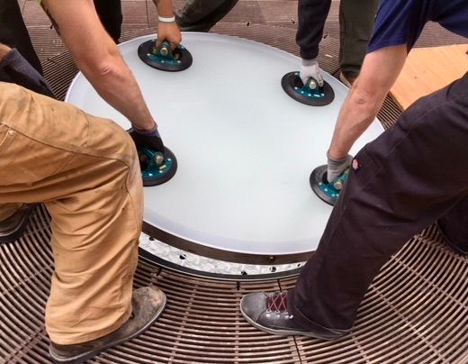 Four people use suction cup handles to lift a large, round, frosted glass panel over a circular metal grate.