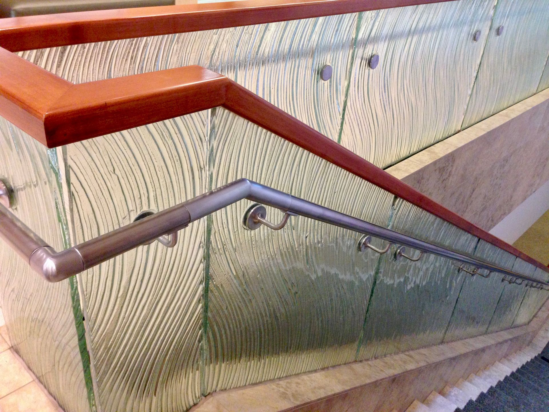 A wooden handrail atop a textured glass partition with a secondary metallic railing mounted to the side for support.