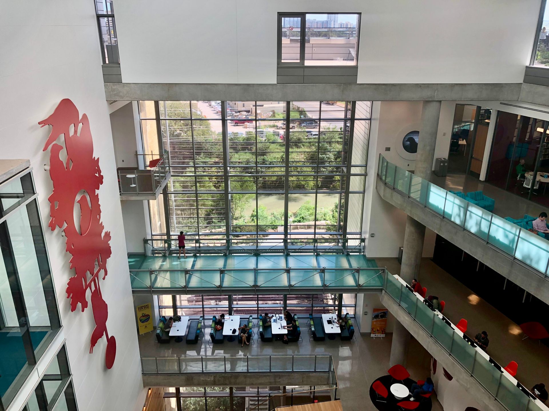 Interior of a multi-story building with a large glass window overlooking a green area; people sit at tables.