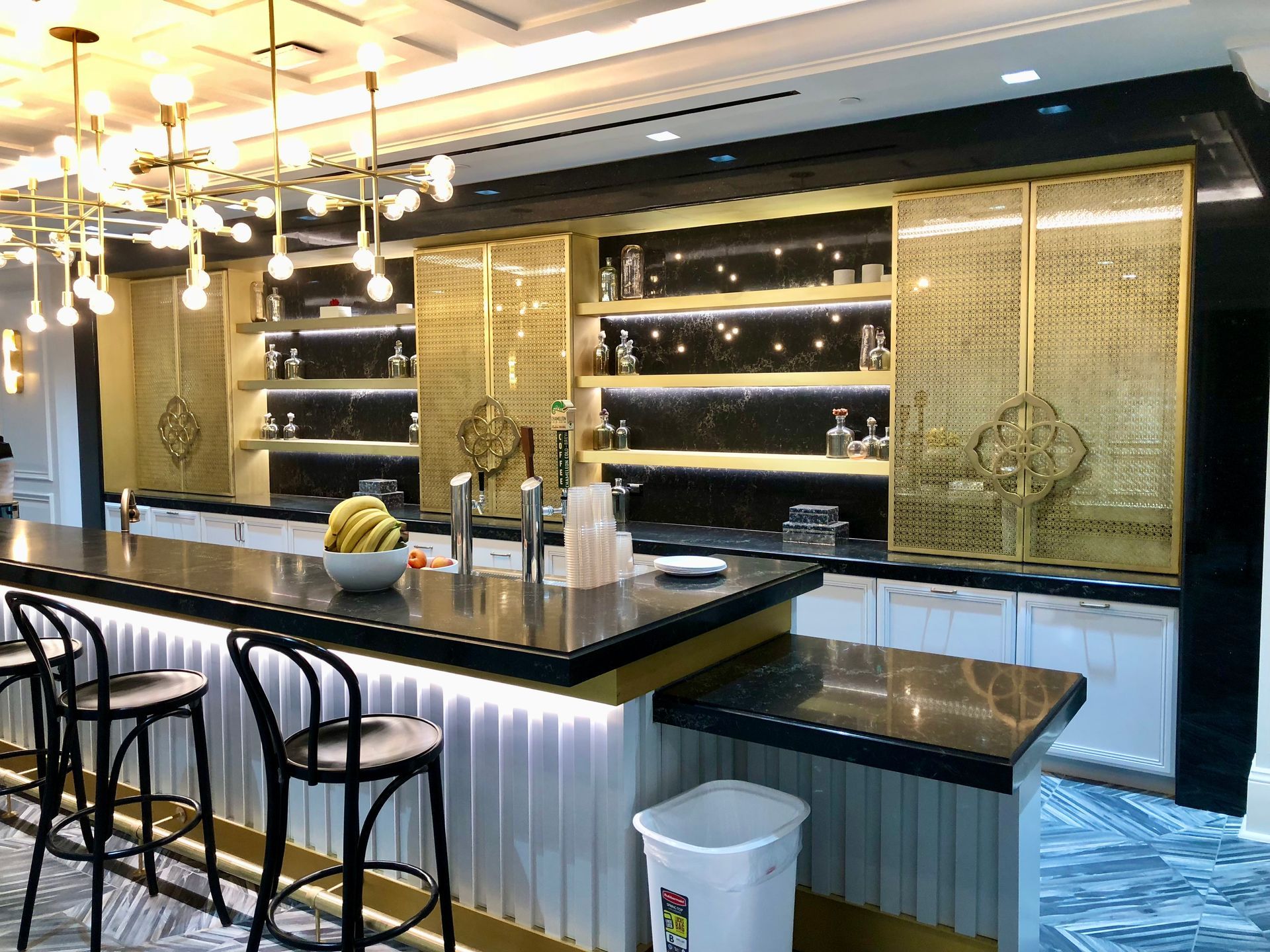 Upscale home bar with gold accents, black countertop, bar stools, and liquor shelves.