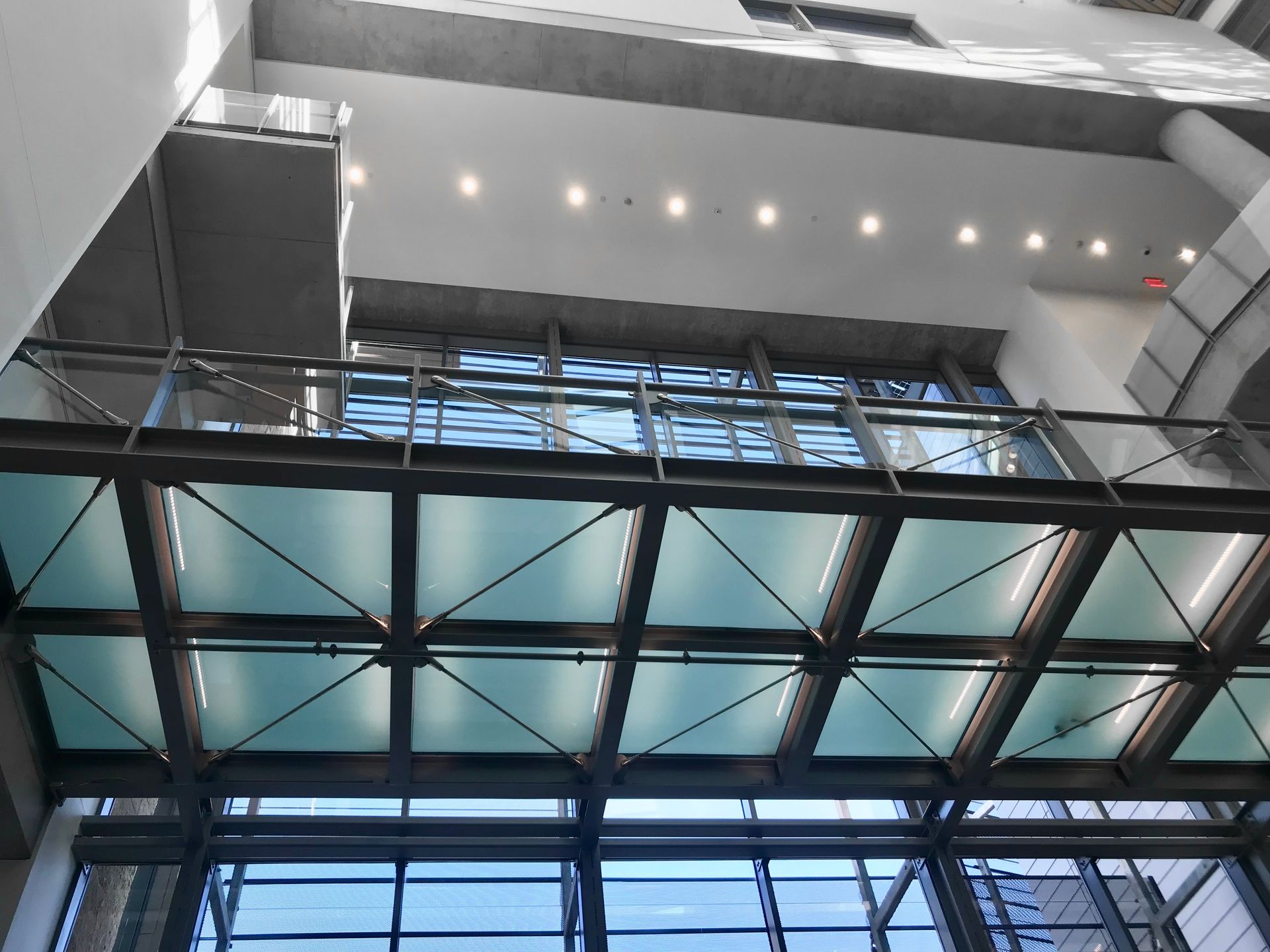Glass walkway inside a building, viewed from below. Metal framework supports the transparent floor panels.