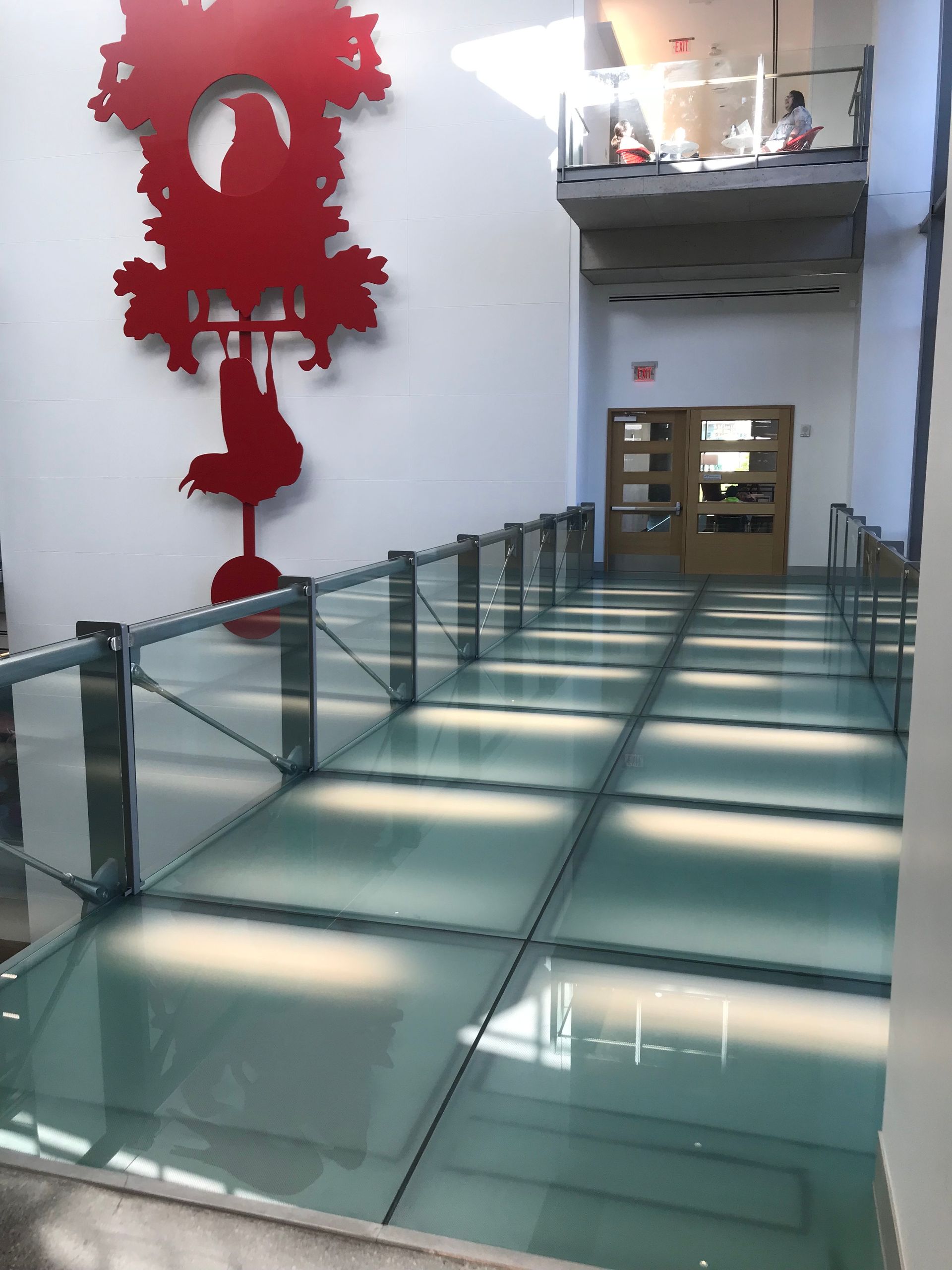 A glass walkway in a building, with a red clock art piece on the wall.