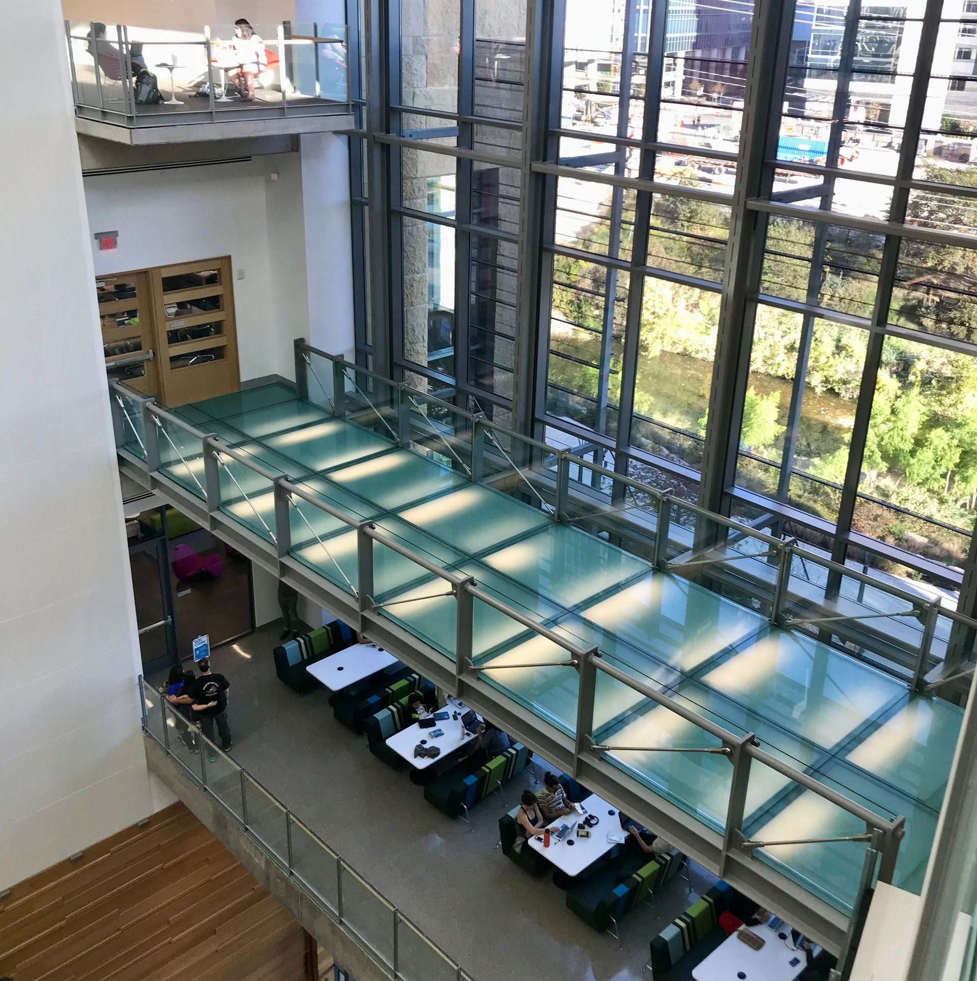 An elevated glass walkway crosses a bright, multi-level atrium with a dining area below and floor-to-ceiling windows.