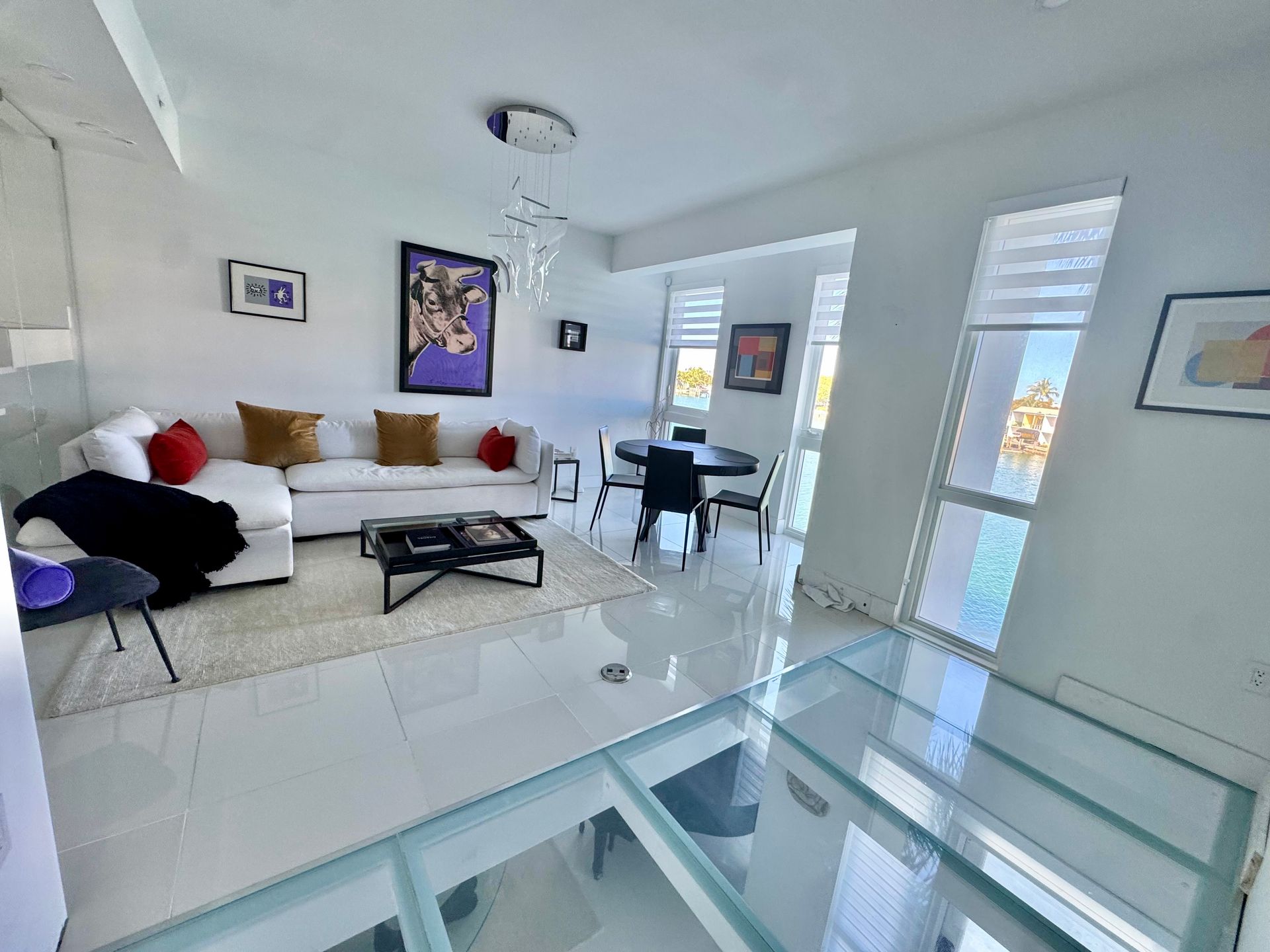 A bright living room features a white sectional sofa, a black dining set, and a glass floor area.