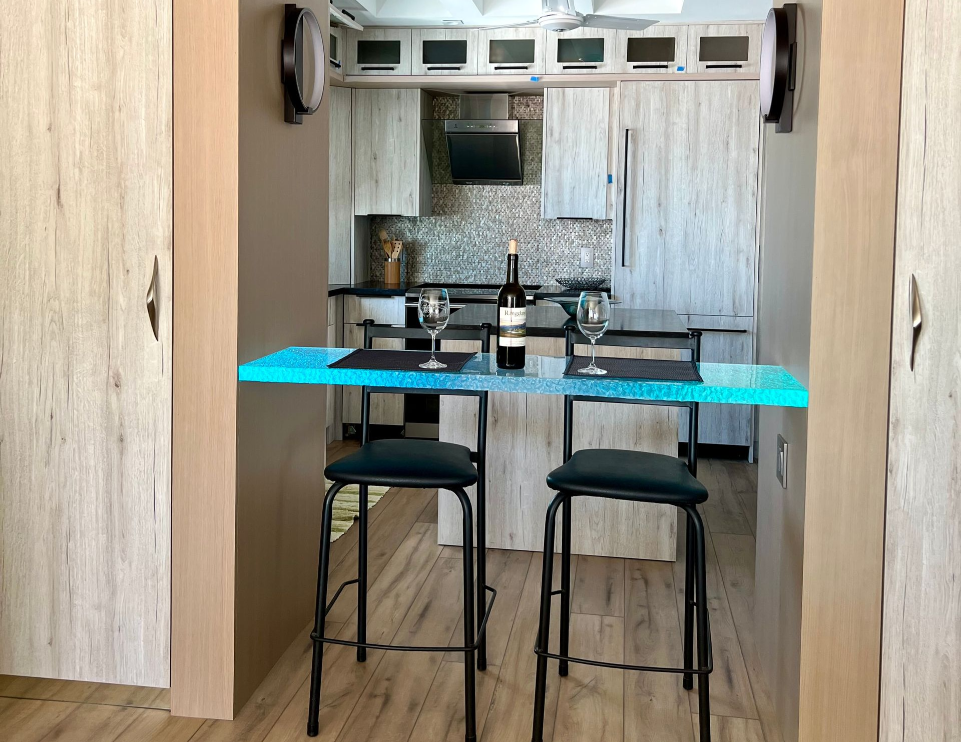 A modern kitchen with a bar table, stools, and wine glasses; viewed from between two light wooden doors.