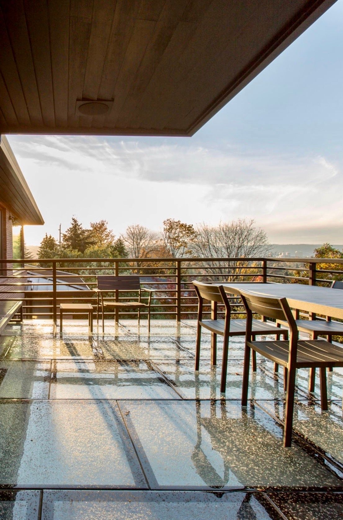 A covered outdoor patio with chairs and a table, overlooking a distant treeline and water under a sunny sky.