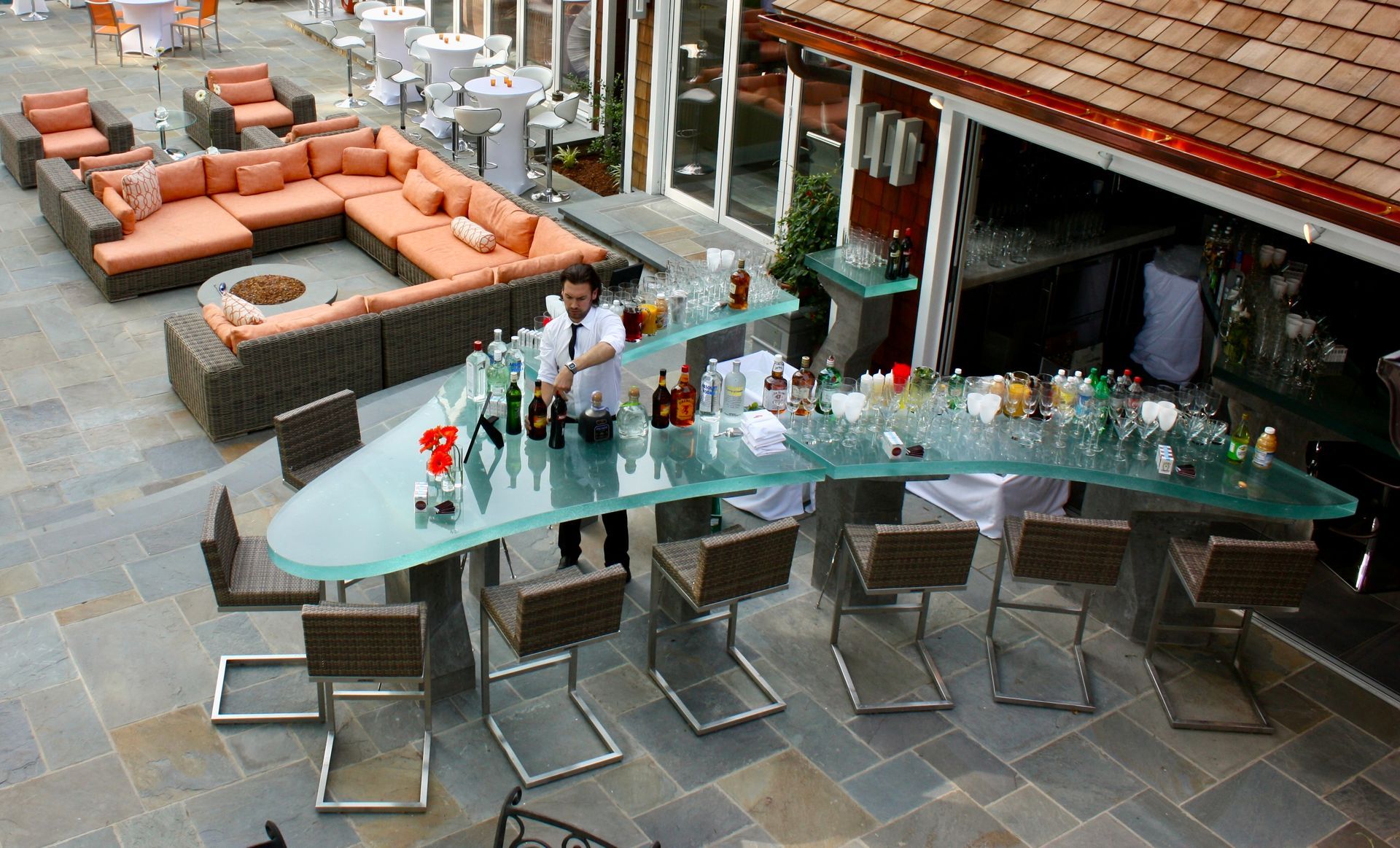 A bartender works behind a modern, curved glass bar on a stone patio with outdoor lounge seating and tables.