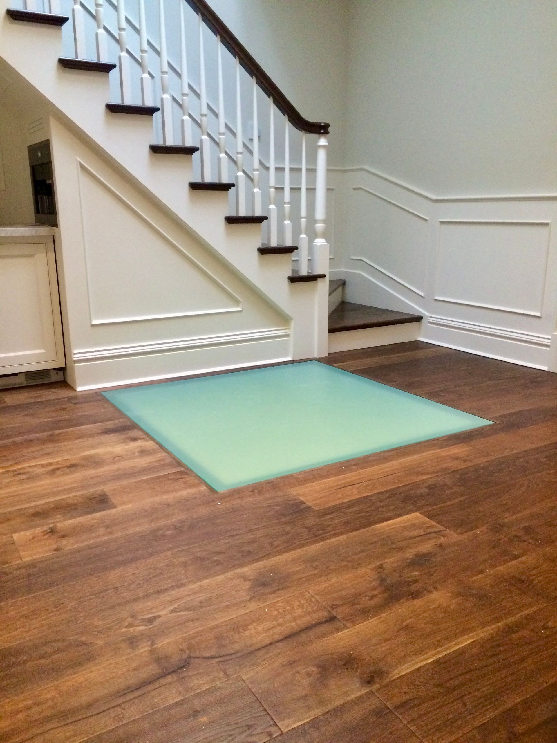 A staircase with white paneling and dark wood steps leads upward, with a large, light-green square patch on the floor.