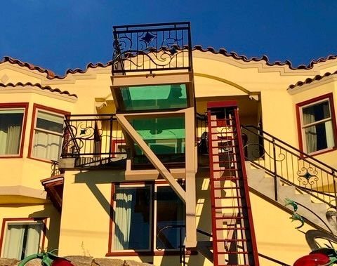 Yellow house exterior with a metal balcony featuring a glass floor and an attached red ladder leading to the ground.