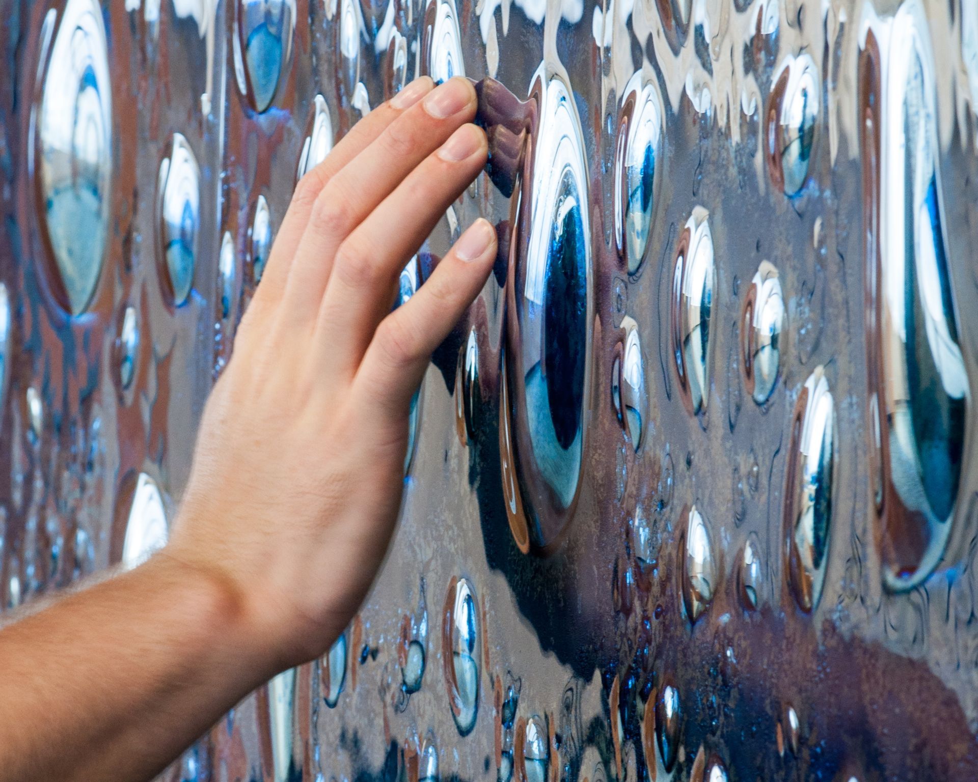 Hand touching a textured, reflective metal wall with distorted droplet shapes.