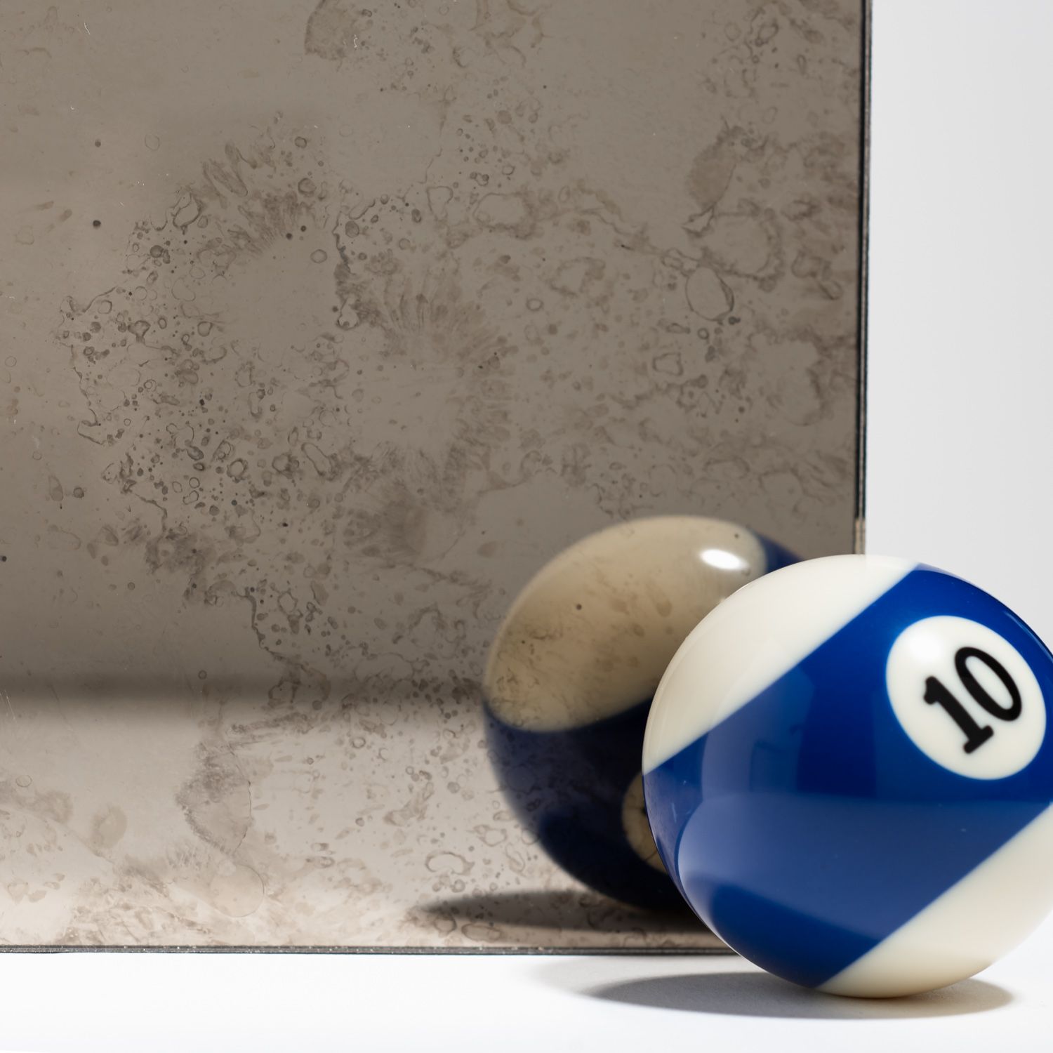 A blue and white number 10 billiard ball sits next to its reflection in a piece of antiqued, distressed mirror glass.