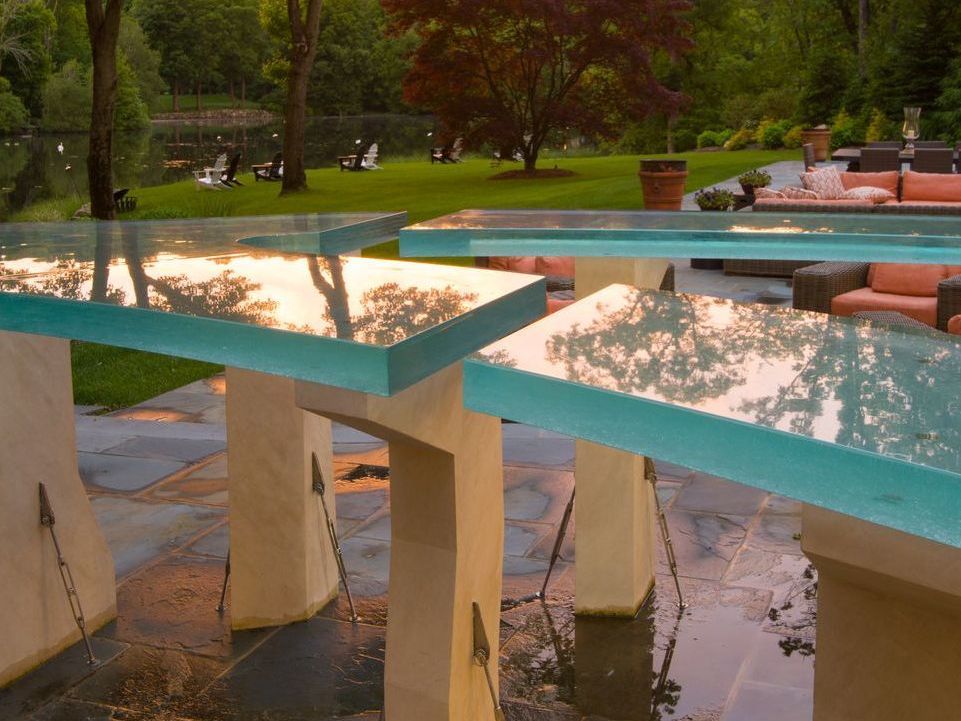 Outdoor bar with blue-green glass tops, white stone bases, and a serene backyard view.