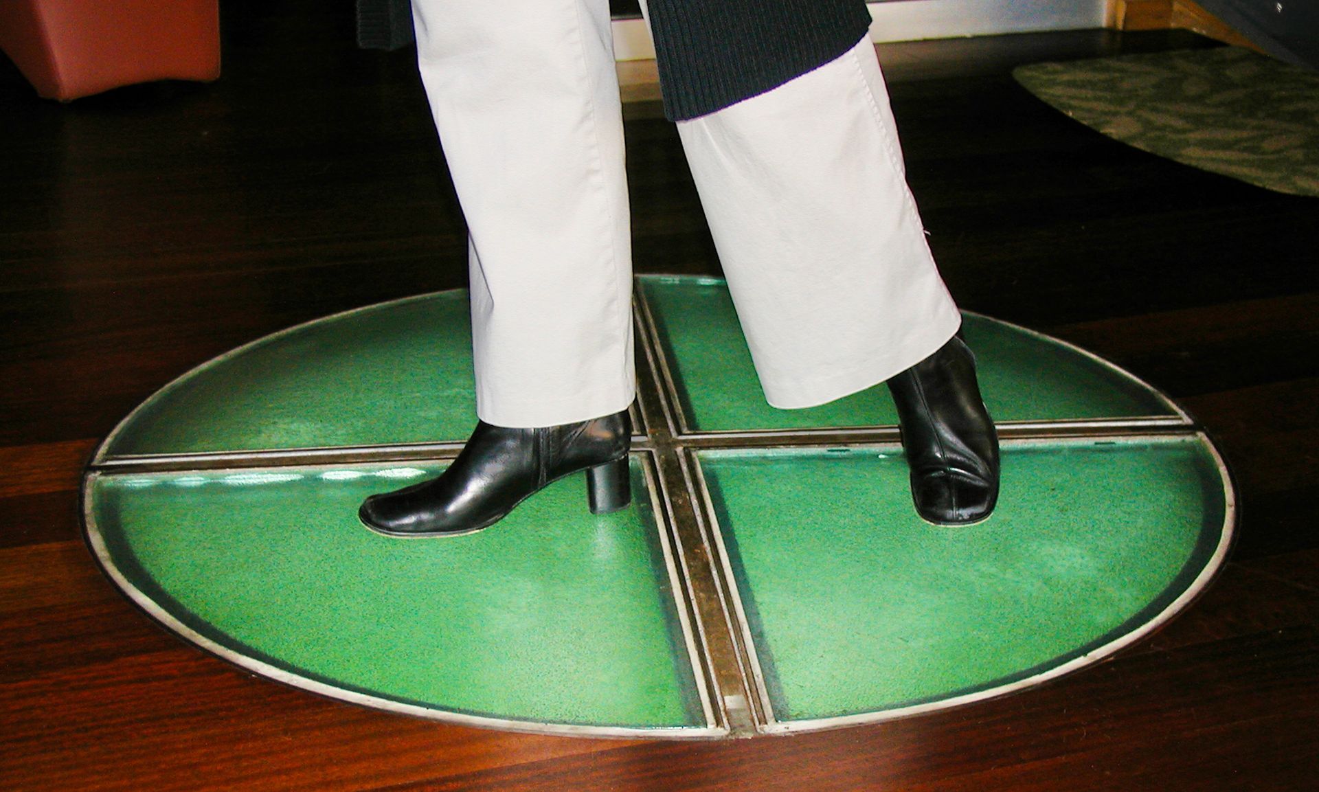 A person stands on a circular, green, segmented glass floor inlay with a metal cross divider set into a wooden floor.