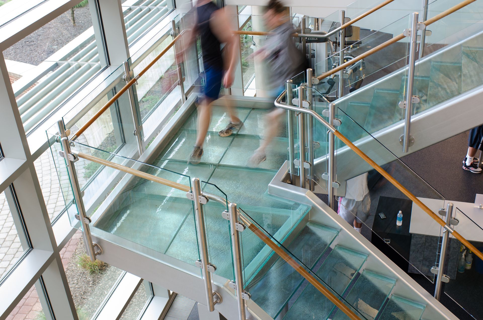 People walking on a glass staircase with steel railings, inside a building with large windows.