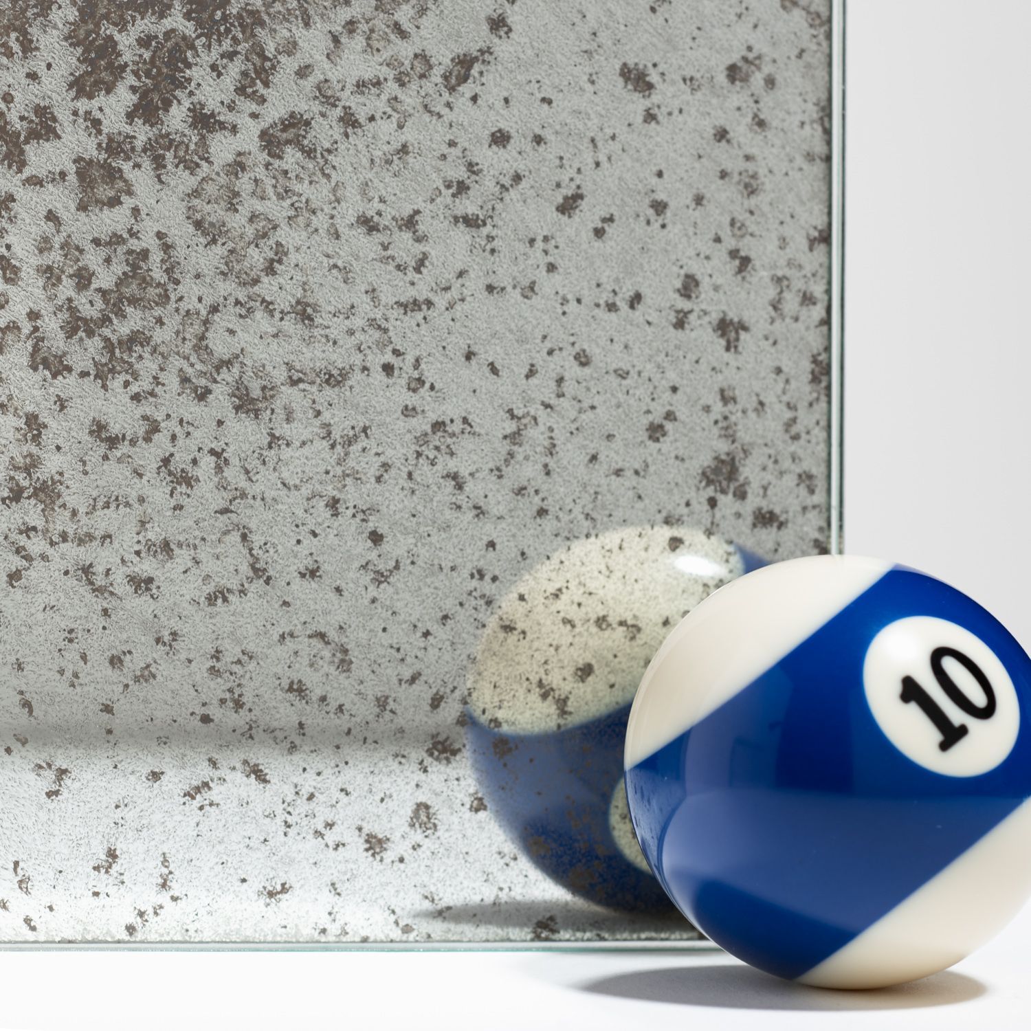 A blue and white number 10 billiard ball rests in front of a piece of speckled, vintage-style antique mirror glass.
