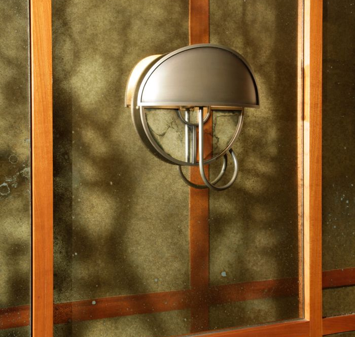 A metal outdoor wall lamp with a dome-shaped shade mounted on an antique mirror textured surface.