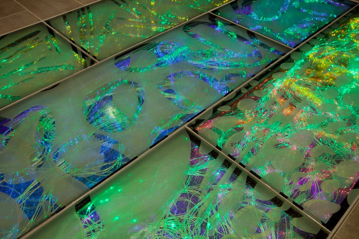 Rectangular glass floor panels illuminated with vibrant green and blue light, featuring swirling, abstract patterns.