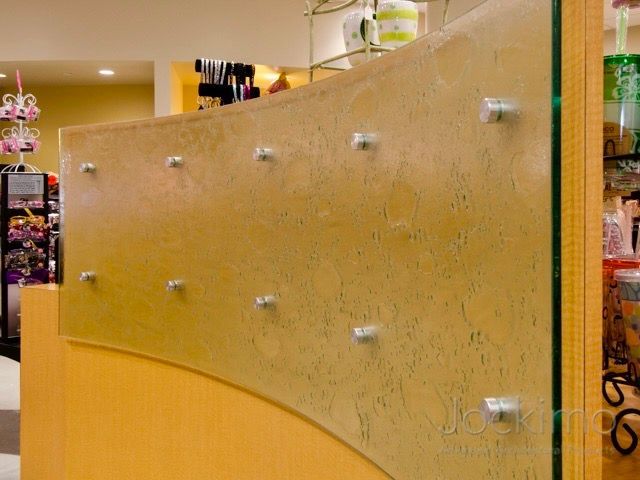 Curved display wall with gold textured glass, clear supports, and tan wood base in a store.