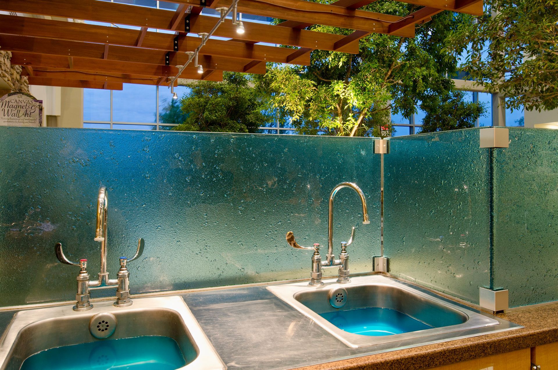 Two stainless steel sinks with chrome faucets set against a textured, teal-colored textured glass backsplash.
