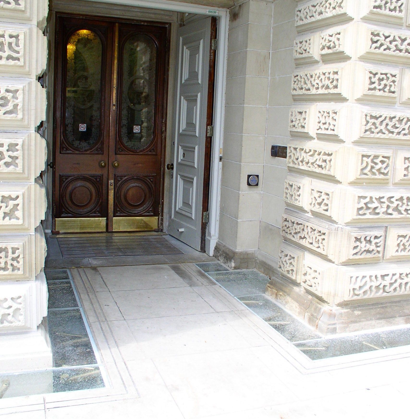 Wooden double doors partially open to a building's entrance, with light-colored stone walls.