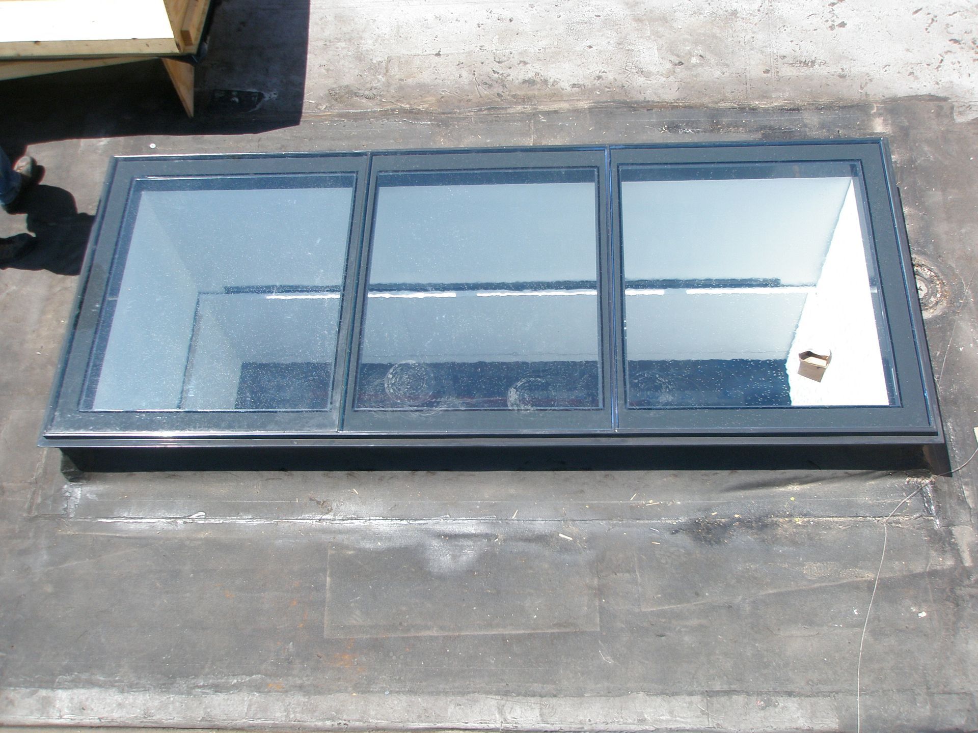 Skylight on a flat, dark roof. The skylight is a rectangle with three glass panels set in a gray frame.