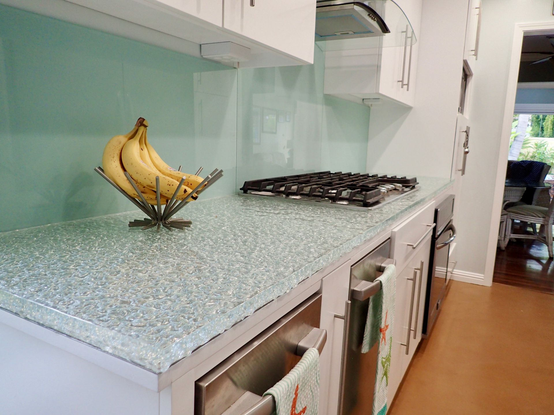 A kitchen counter with a light blue glass backsplash, a glass-flecked countertop, a bowl of bananas, and a gas cooktop.