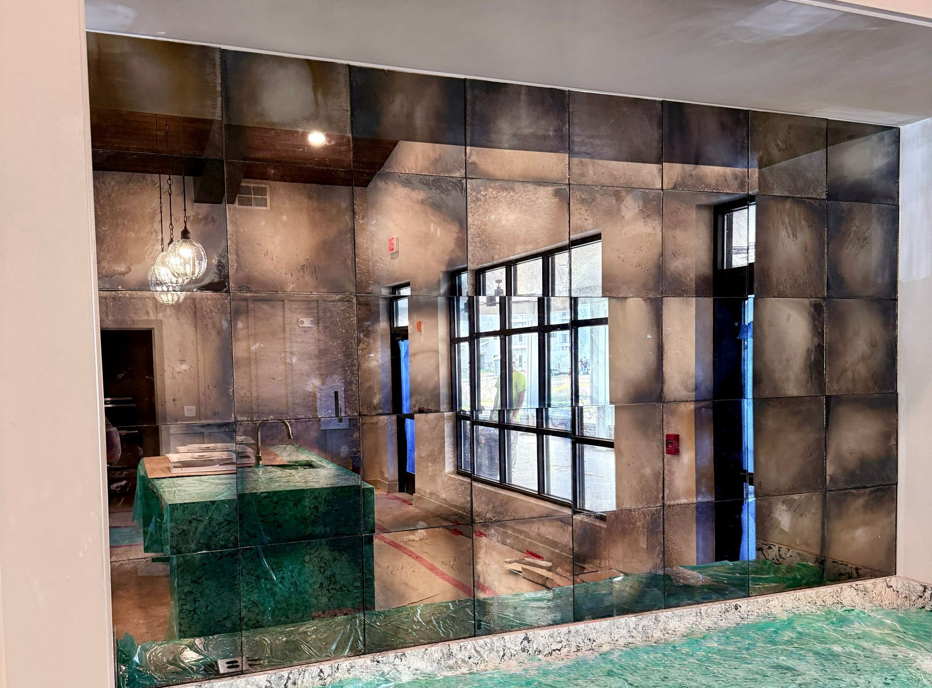 An antique-style mirror wall composed of square panels reflects an interior room with a green counter and large windows.