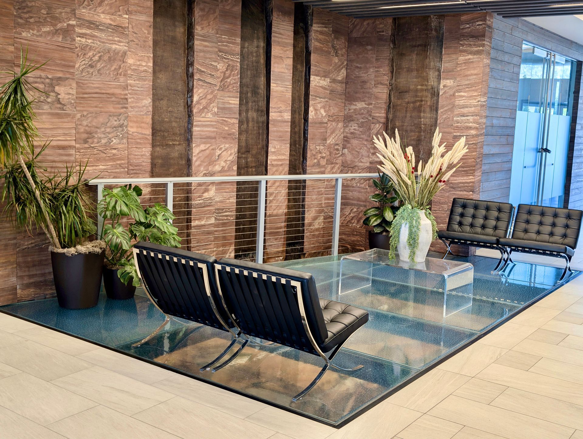 Modern lobby featuring black Barcelona-style chairs on a glass floor with brown stone walls and potted plants.
