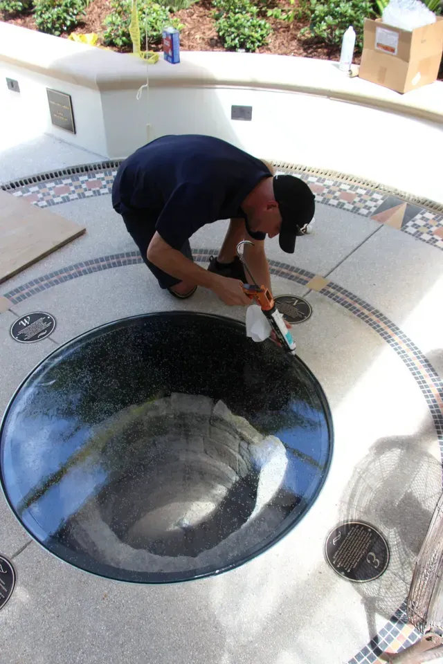 A worker in a dark shirt kneels on a stone plaza, applying caulk to the edge of a circular glass feature.