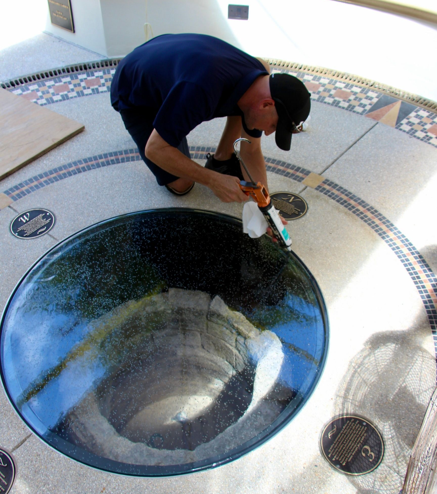 A person in a dark shirt and cap uses a sealant gun to seal the edge of a glass floor panel overlooking a stone well.