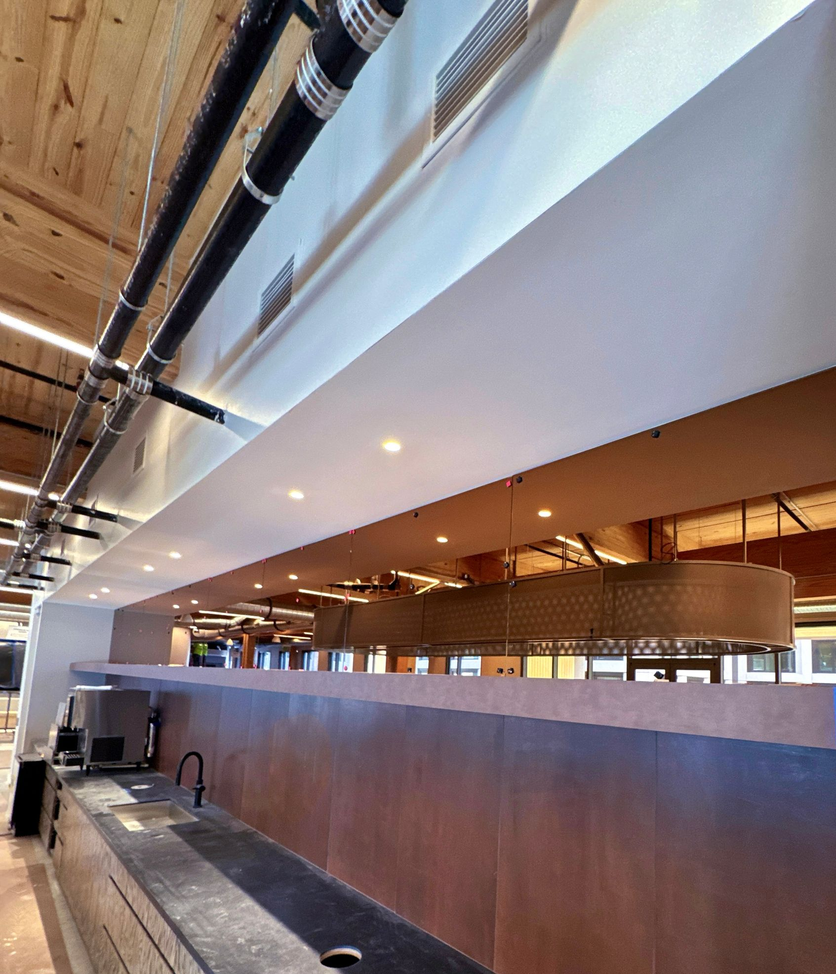 Bar interior with copper and cream-colored surfaces, black pipes overhead, and recessed lighting.