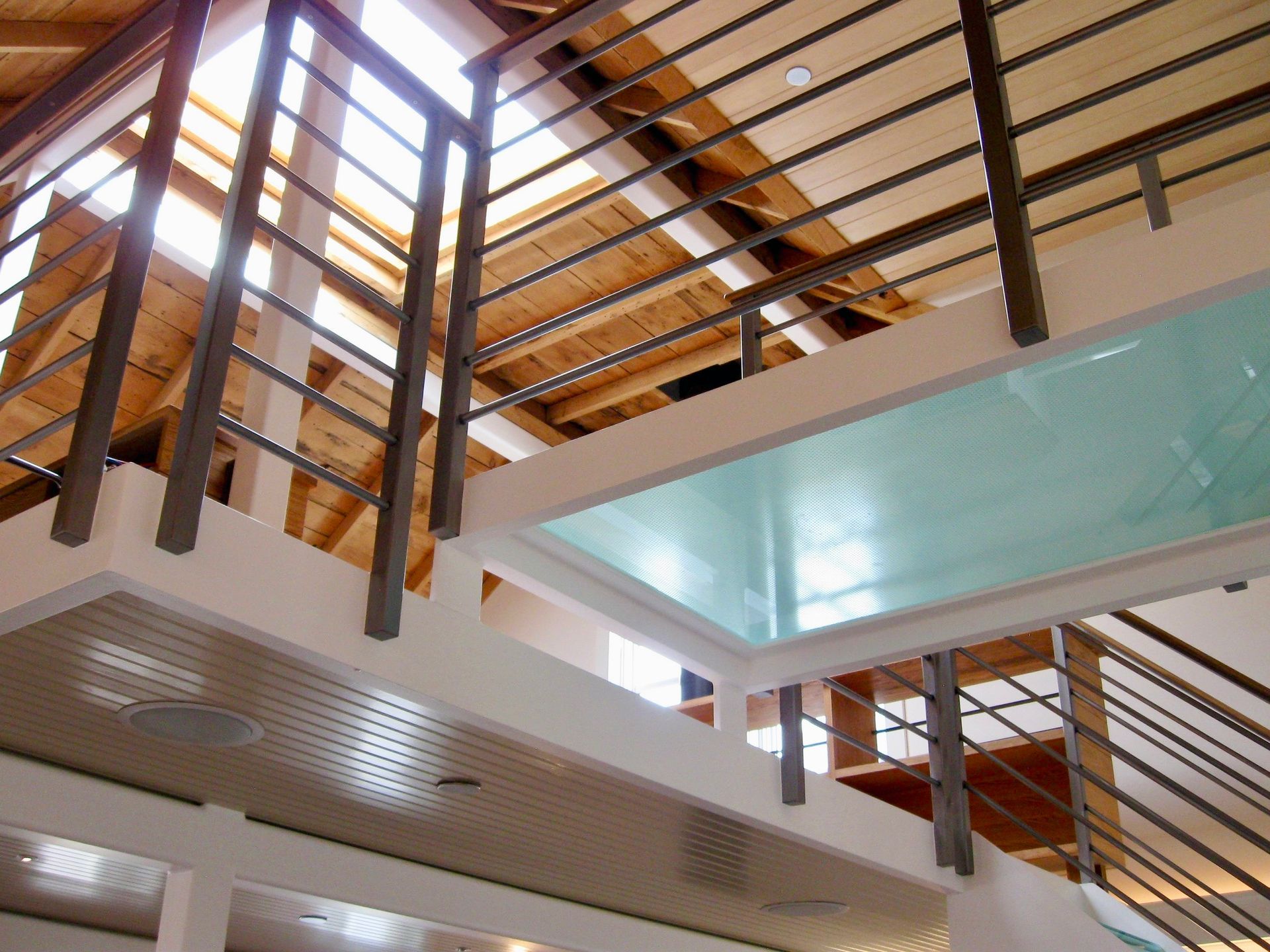 A modern home interior features a raised wooden platform, a glass floor section, and metal railings under a bright skylight.